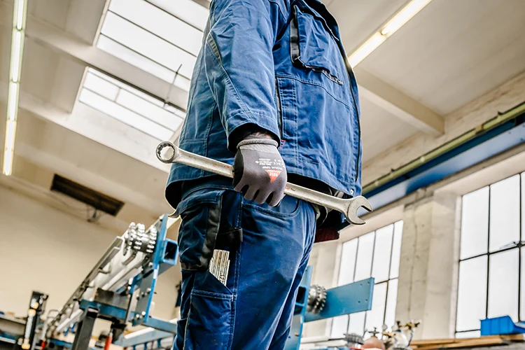 Mitarbeiter in blauer Uniform und schwarzen Handschuhen hält einen großen Schraubenschlüssel in der Hand und steht in einer Industrieanlage mit Maschinen im Hintergrund. Der Raum ist durch Deckenleuchten und große Fenster gut beleuchtet.